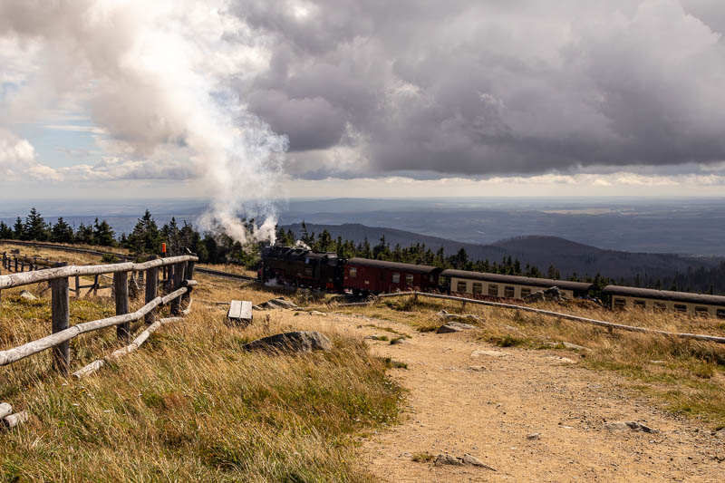 Historische Dampflok der Brockenbahn zieht Waggons um eine Kurve, wobei Rauch aufsteigt. Im Vordergrund sind ein sandiger Wanderweg und ein Holzzaun zu sehen, mit weitem Blick über den Harz im Hintergrund.