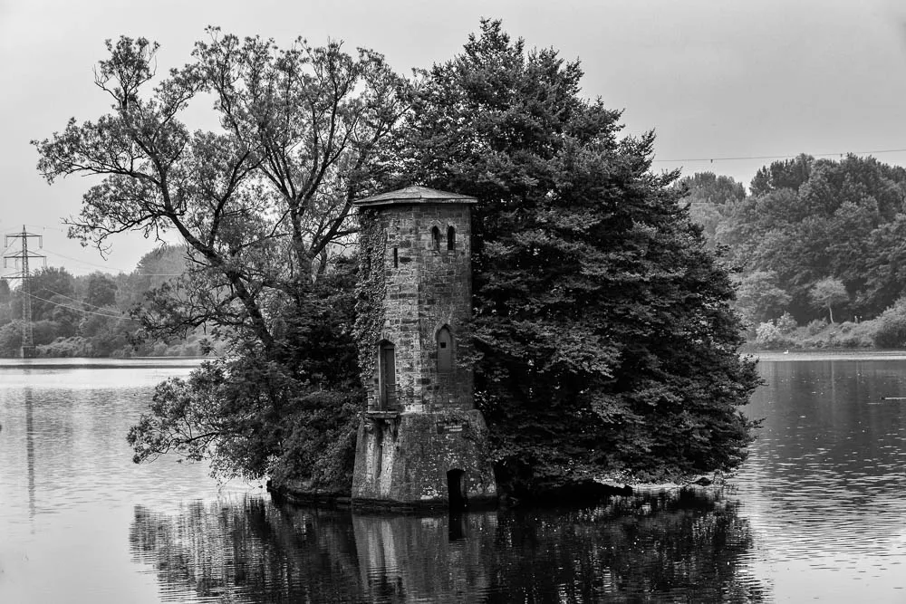 Schwarzweißaufnahme des Mäuseturms im Hengsteysee auf einer kleinen Insel mit Bäumen und Spiegelung im Wasser