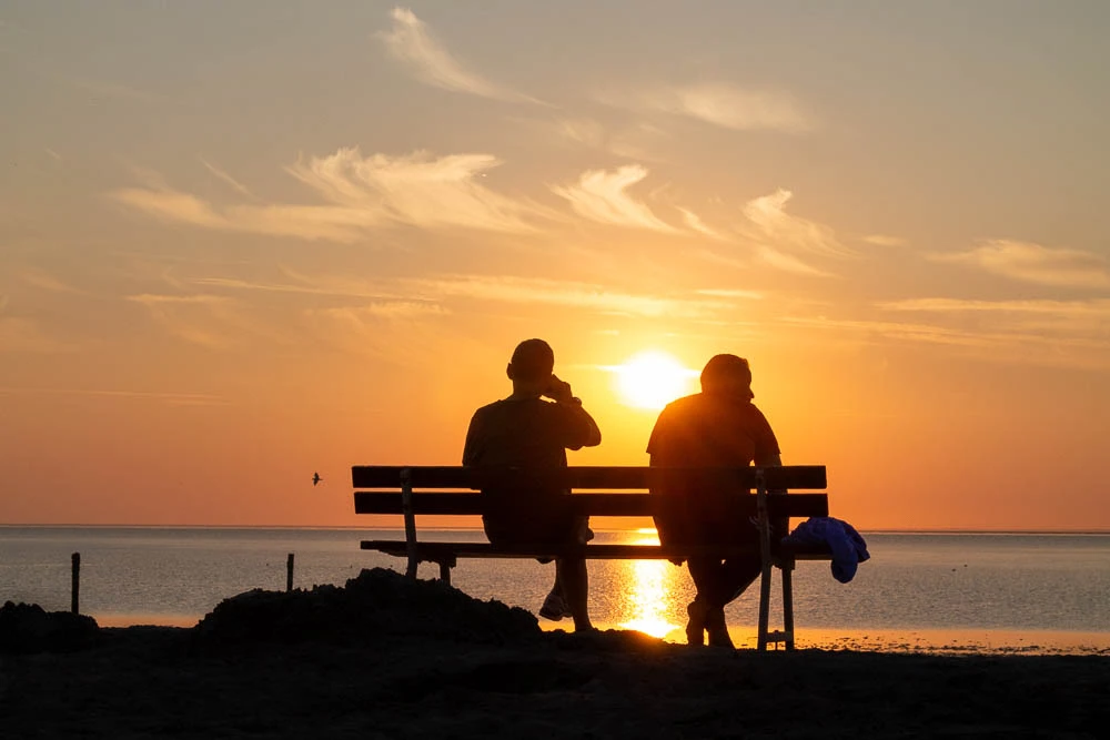 Zwei Personen sitzen auf einer Bank und schauen bei Sonnenuntergang auf die Nordsee in Norddeich.