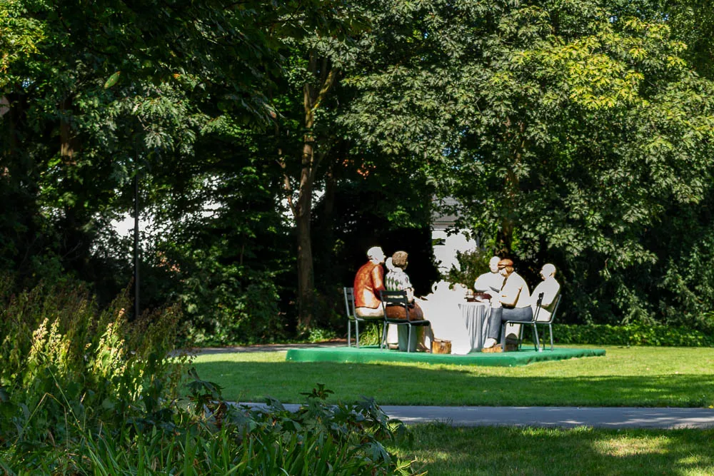 Mehrere Figuren der Ausstellung Alltagsmenschen sitzen an einem gedeckten Tisch in einem Park.