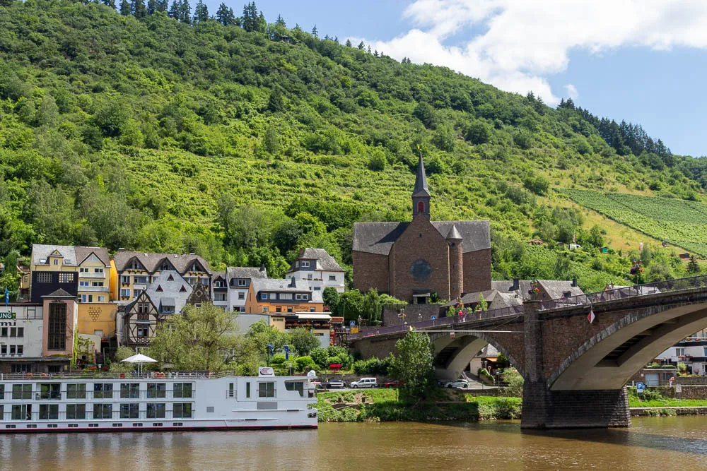 Blick vom Schiff auf Cochem mit Moselbrücke, Kirche und Weinbergen am Hang.