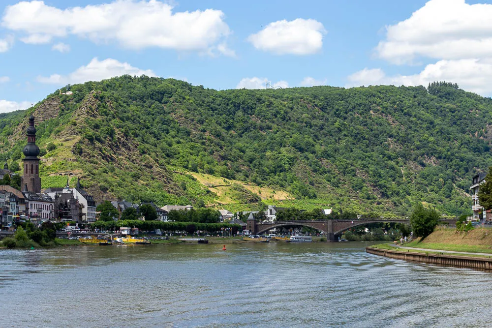 Blick vom Schiff auf die Mosel mit Brücke, Weinbergen und einem Ort am Flussufer.