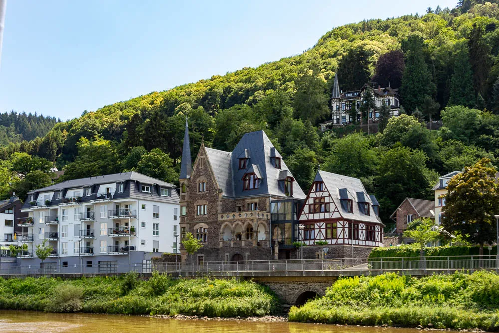 Historische Villa und Fachwerkhaus am Moselufer vor bewaldetem Hang.