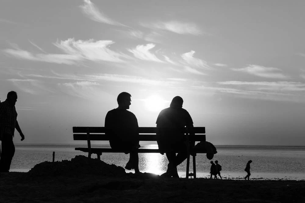 Silhouetten von Menschen auf einer Bank und am Strand vor dem hellen Horizont an der Nordsee.