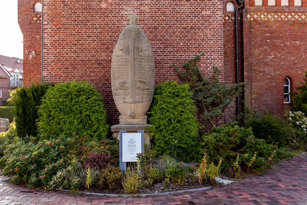 Ein Gedenkstein steht vor der Backsteinfassade der Inselkirche Langeoog in einem bepflanzten Vorgarten.