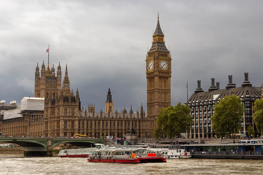 Der Palace of Westminster mit dem Elizabeth Tower und der Big-Ben-Uhr am Ufer der Themse, davor fahren Ausflugsschiffe auf dem Fluss.