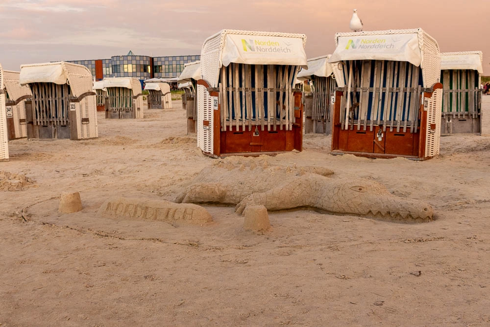 Aus Sand geformtes Krokodil am Strand von Norddeich vor Strandkörben.